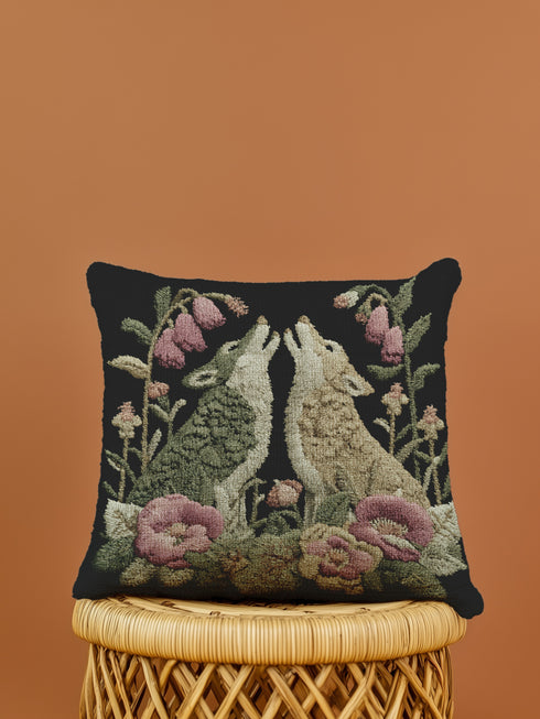 A square woven tapestry pillow with two howling wolves on a black background. The wolves are surrounded by pink and green botanical flowers, creating a vintage, folkloric scene. This decorative cushion is perfect for cottagecore, dark academia, and gothic home decor.